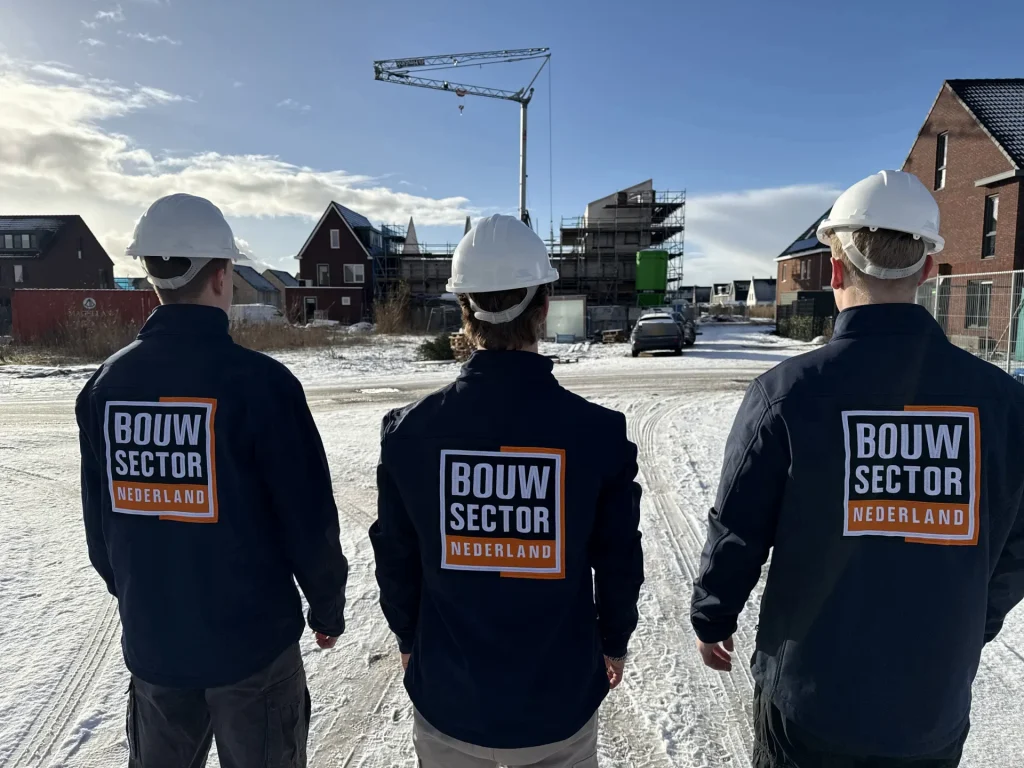 Drie vakmensen van Bouwsector Nederland in donkerblauwe jassen en witte helm kijken naar een nieuwbouwwijk